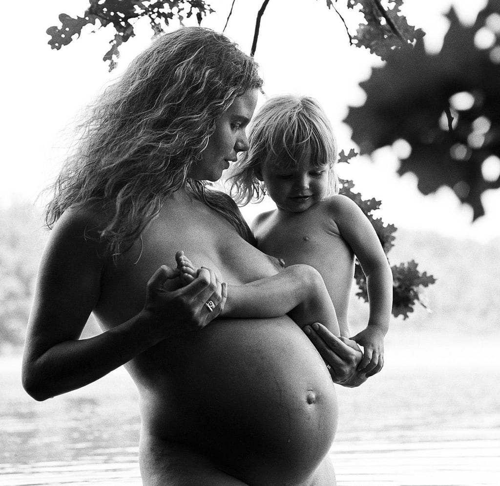 Schwangere steht im Wasser und hält ihr Kleinkind auf dem Arm. Gemeinsam schauen sie auf den Babybauch beim Babybauch Shooting in Hamburg in der Natur am See