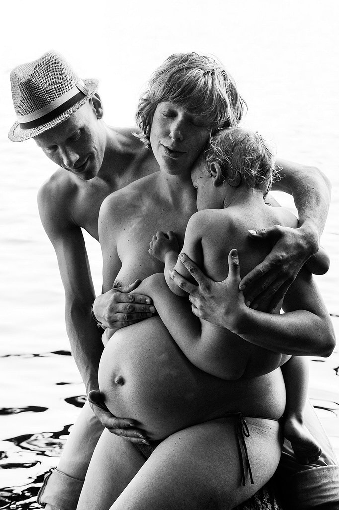 Mama und Papa sitzen gemeinsam mit ihrem Kind im Arm am See beim Babybauch Shooting in Hamburg in der Natur