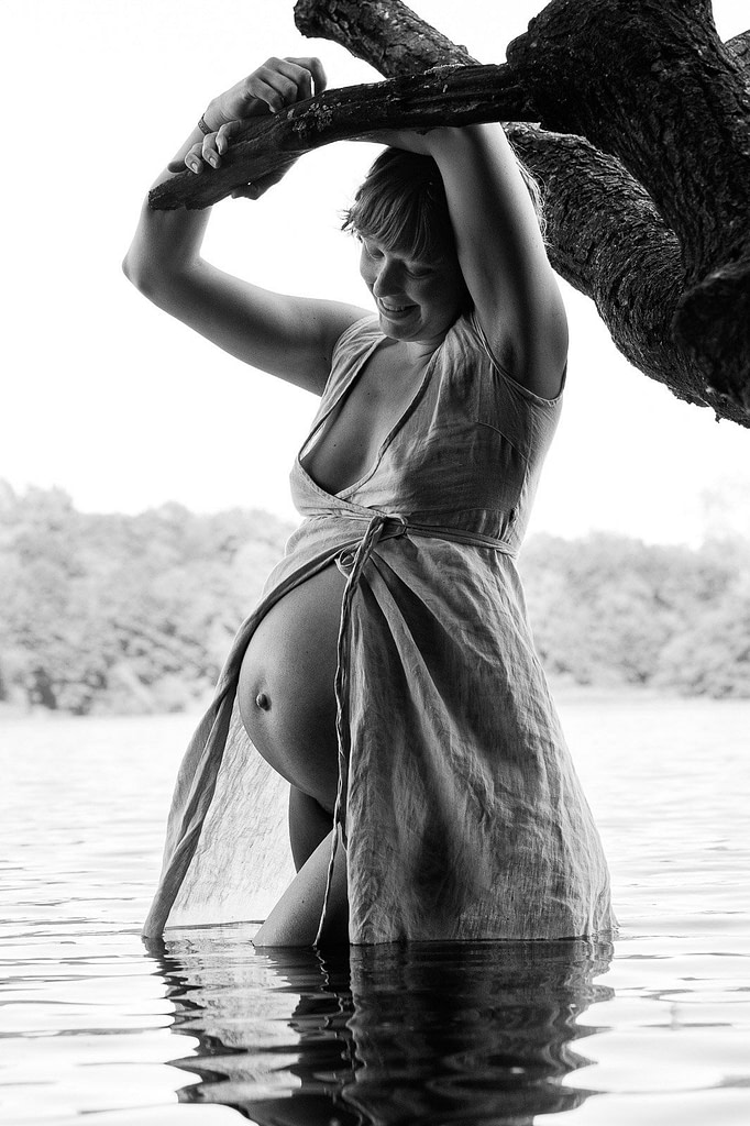 Schwangere steht im Kleid im Wasser und schaut auf ihren Babybauch beim Schwangerschaftsfotoshooting in Hamburg am See in der Natur