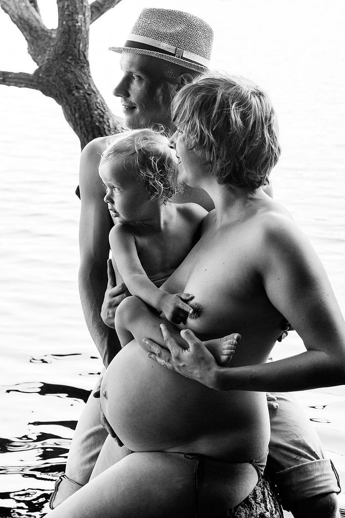 Mama und Papa sitzen gemeinsam mit ihrem Kind im Arm am See und schauen auf das Wasser beim Babybauch Shooting in Hamburg in der Natur