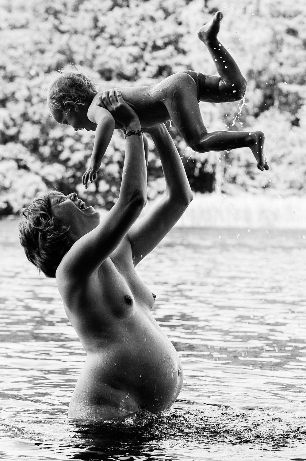 Mama steht im Wasser und hebt ihr Kind mit Schwung hoch in die Luft beim Babybauch Shooting in Hamburg am See