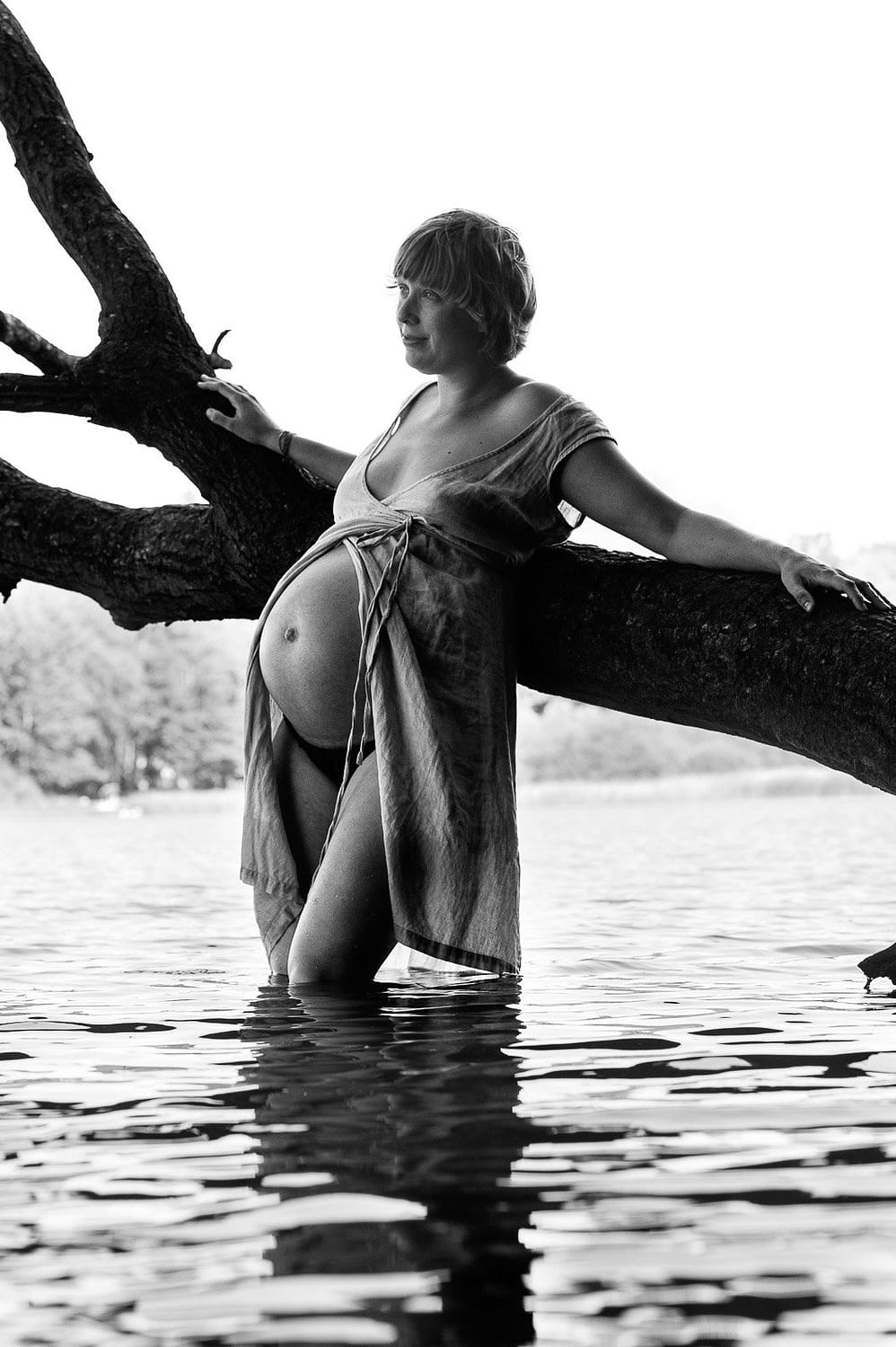 Schwangere steht im Kleid im Wasser am Baum gelehnt beim Babybauch Shooting in Hamburg am See in der Natur